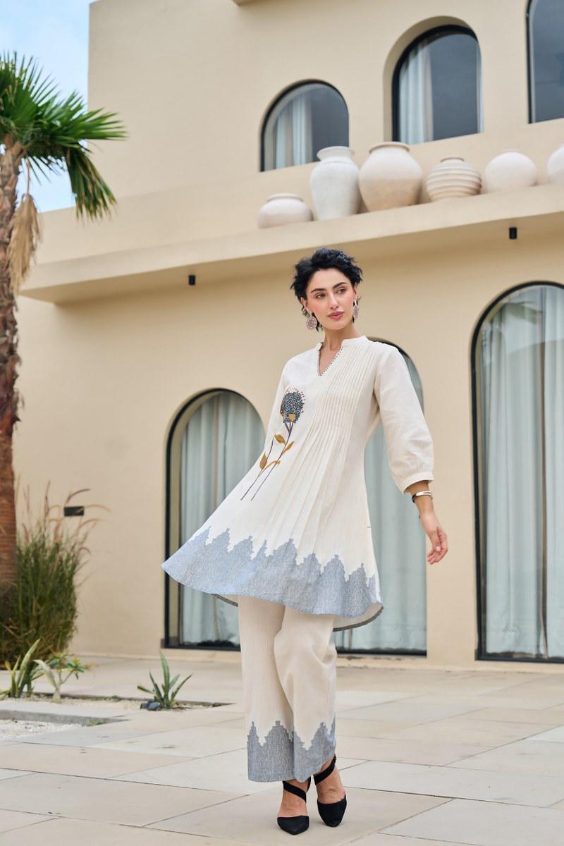 Woman in a white and gray traditional outfit standing in front of a modern building with decorative pots.