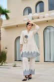Woman in a cream and gray traditional outfit standing in front of a building with decorative elements.