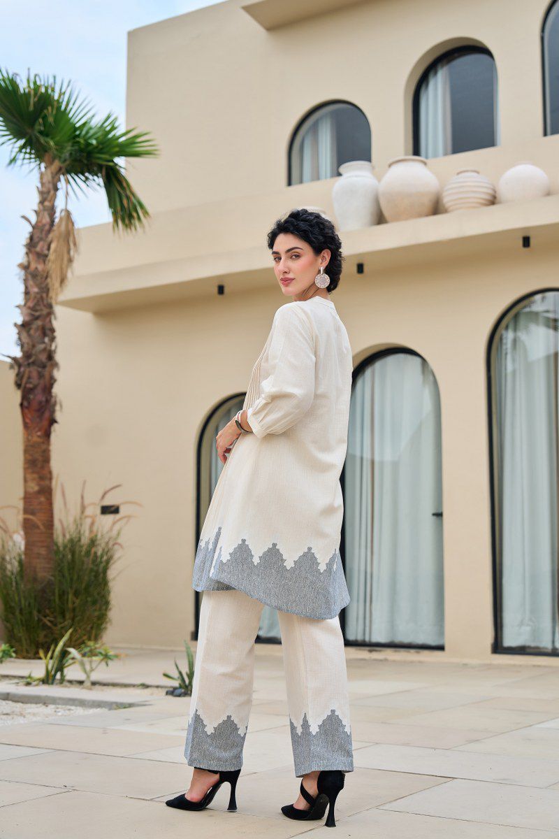 Woman in a white and gray traditional outfit standing in front of a building with decorative elements.